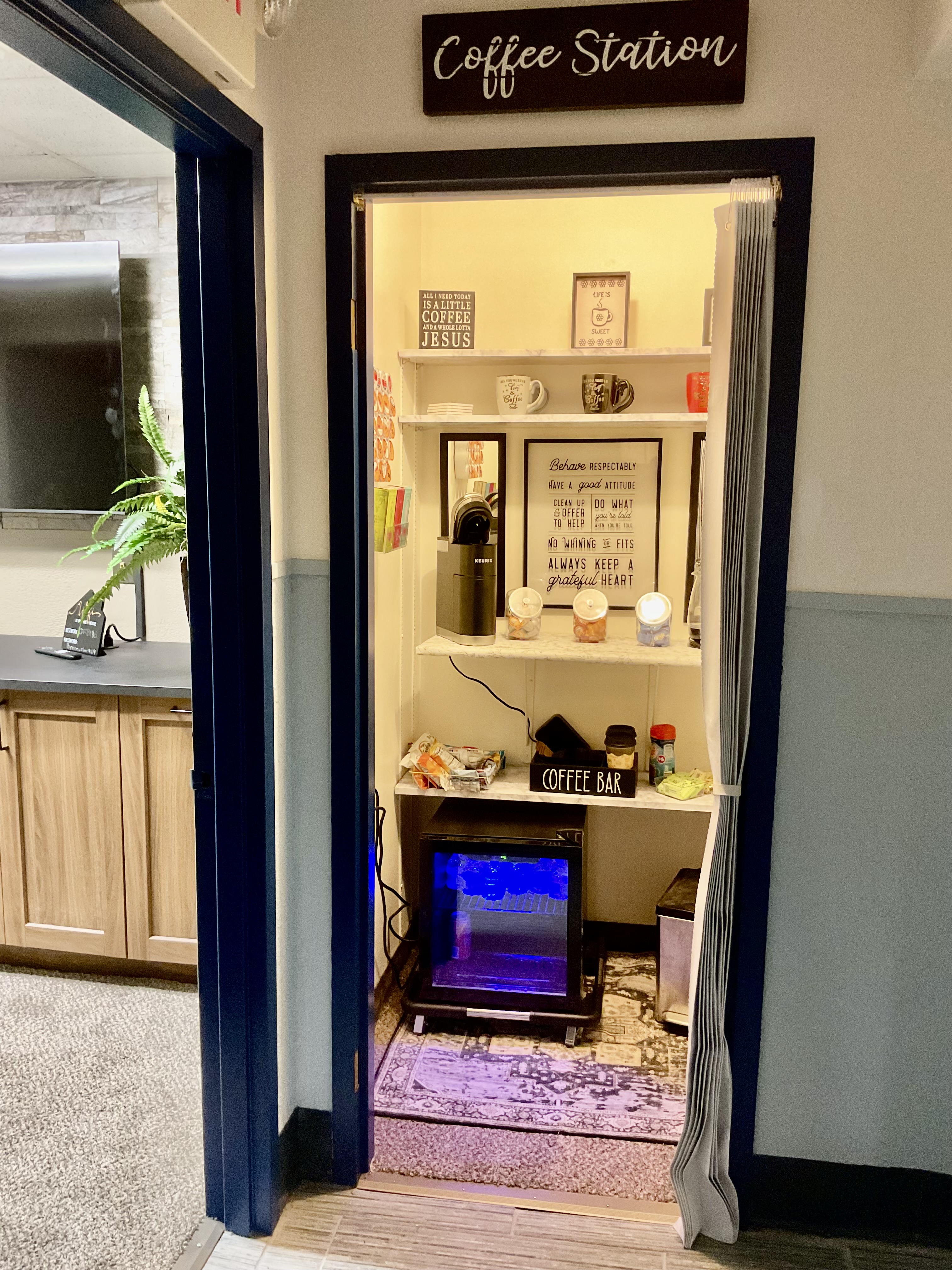 Navy-framed coffee station alcove with a hand-lettered sign, mugs on wooden shelves, a brewer, and a refrigerator glowing below